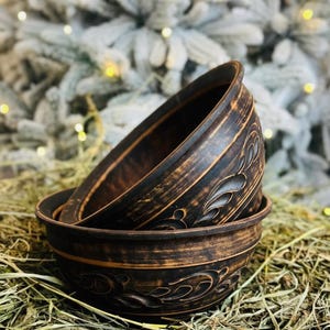 Puede incluir: Tres cuencos de cerámica marrón oscuro apilados con patrones florales decorativos. Los cuencos tienen un aspecto rústico y envejecido, con una superficie texturizada. Los cuencos están colocados sobre un lecho de heno, con un fondo borroso de un árbol de hoja perenne nevado.