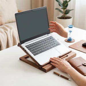 May include: A silver laptop computer resting on a dark brown wooden stand. The stand features an adjustable angled design with a horizontal support bar. The laptop has a black keyboard and a dark screen, positioned on a white desk surface.