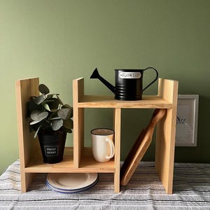 Pode incluir: Uma prateleira de madeira com vários compartimentos exibe um regador preto, uma caneca branca, uma pá de madeira e uma planta em vaso com a etiqueta "FRESH HERBS". Um prato branco com borda azul também é visível.