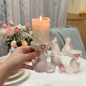 Może przedstawiać: Biały ceramiczny świecznik w kształcie leżącego królika z zamkniętymi oczami, ozdobiony różową różą na krawędzi. W świeczniku znajduje się zapalona świeca pieńkowa z topniejącym woskiem, trzymana na tle kwiatów.