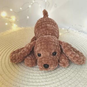 May include: A plush, brown crocheted dog toy with floppy ears and a curled tail. The dog has black eyes and a black nose. The toy is lying on a textured, off-white surface.