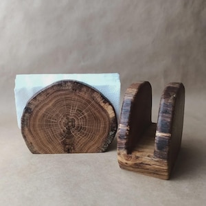 May include: Two wooden napkin holders. One is a half-circle shape with a cross-section of a tree trunk, holding white napkins. The other is a rectangular shape with a U-shaped opening. Both are made of brown wood.