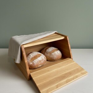 Könnte beinhalten: Eine Brotdose aus Holz mit einer Klappe, die zwei runde Brotlaibe enthält. Ein Leinentuch bedeckt teilweise das obere Regal. Die Dose ist aus hellem Holz gefertigt und steht auf einer weißen Oberfläche.