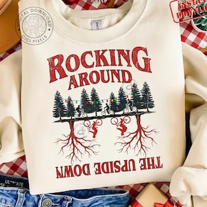 Może przedstawiać: Kremowa bluza z czerwonym napisem "ROCKING AROUND THE UPSIDE DOWN". Grafika przedstawia rowerzystów jadących przez las choinek. Projekt zawiera korzenie drzew i sznur światełek.