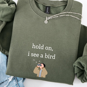May include: An olive green sweatshirt with the text "hold on, i see a bird" and an illustration of a person with binoculars. A silver necklace and blue jeans are also in the image.