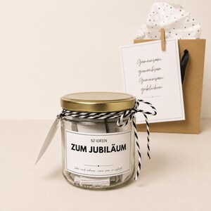 May include: A clear glass jar with a gold lid, filled with paper slips. The label reads 52 IDEEN ZUM JUBILÄUM. It is tied with black and white baker's twine. Behind it is a brown paper gift bag with a white card featuring the text Gemeinsame.