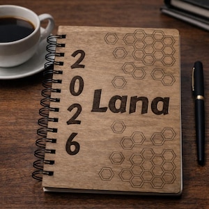 Könnte beinhalten: Ein holzgebundenes Notizbuch mit dem Namen "Lana" und der Jahreszahl "2026" eingraviert. Ein schwarzer Stift und eine Tasse Kaffee liegen auf einer Holzoberfläche. Das Notizbuch hat eine Spiralbindung und ein Wabenmuster.