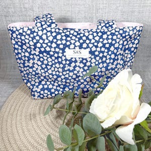 Könnte beinhalten: Eine marineblaue Tragetasche mit weißem Blumenmuster und rosa Futter. Die Tasche hat zwei Griffe und ein Etikett mit der Aufschrift "S&S Shuttle and Stitch". Eine weiße Rose und Grünzeug sind im Vordergrund.