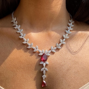 Puede incluir: Un conjunto de collar y pendientes de plata. El collar presenta un diseño de hojas con piedras transparentes y un gran colgante rojo en forma de lágrima. Los pendientes tienen un diseño similar con una gema roja.