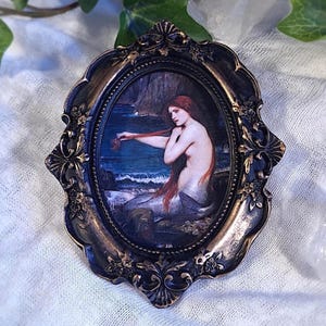 Op de afbeelding: Ovaal ingelijste kunstdruk met een zeemeermin met lang rood haar, zittend bij de oceaan. De lijst heeft een sierlijke, donker bronzen afwerking. Het kunstwerk toont een vrouw met lang rood haar, zittend bij de oceaan.