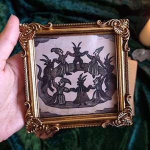 Puede incluir: Un pequeño marco cuadrado dorado con detalles ornamentados contiene una ilustración en blanco y negro de figuras bailando en círculo. La obra de arte tiene una estética vintage. El marco se sostiene en una mano.