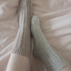 May include: A pair of knitted socks in light gray and sage green, with a cable knit pattern. The socks are on a bed with a pink and white striped sheet.