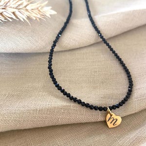 Puede incluir: Un collar de cuentas negras con un colgante de coraz&oacute;n dorado. El colgante de coraz&oacute;n tiene grabada la letra "M". El collar se muestra sobre una tela beige.