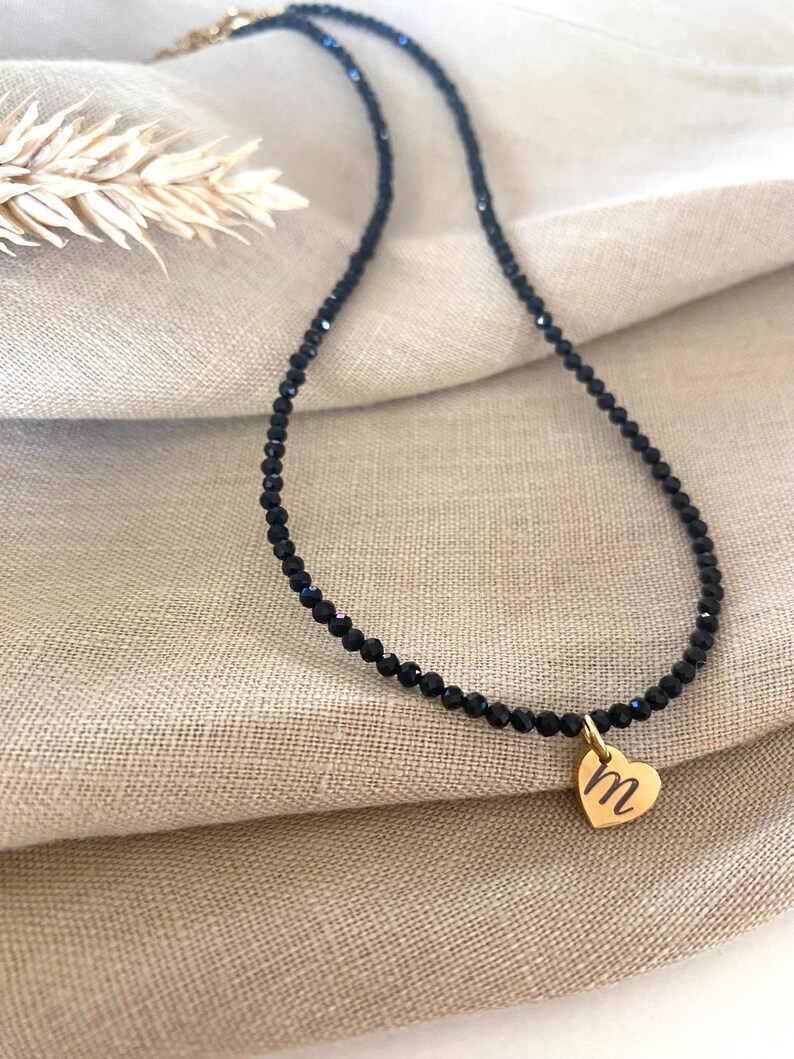 Puede incluir: Un collar de cuentas negras con un colgante de coraz&oacute;n dorado. El colgante de coraz&oacute;n tiene grabada la letra "M". El collar se muestra sobre una tela beige.