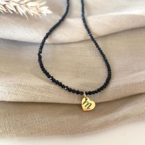 Puede incluir: Un delicado collar de cuentas negras con un colgante de coraz&oacute;n dorado. El colgante de coraz&oacute;n presenta la letra "m" en cursiva. El collar se muestra sobre una tela de tono neutro.