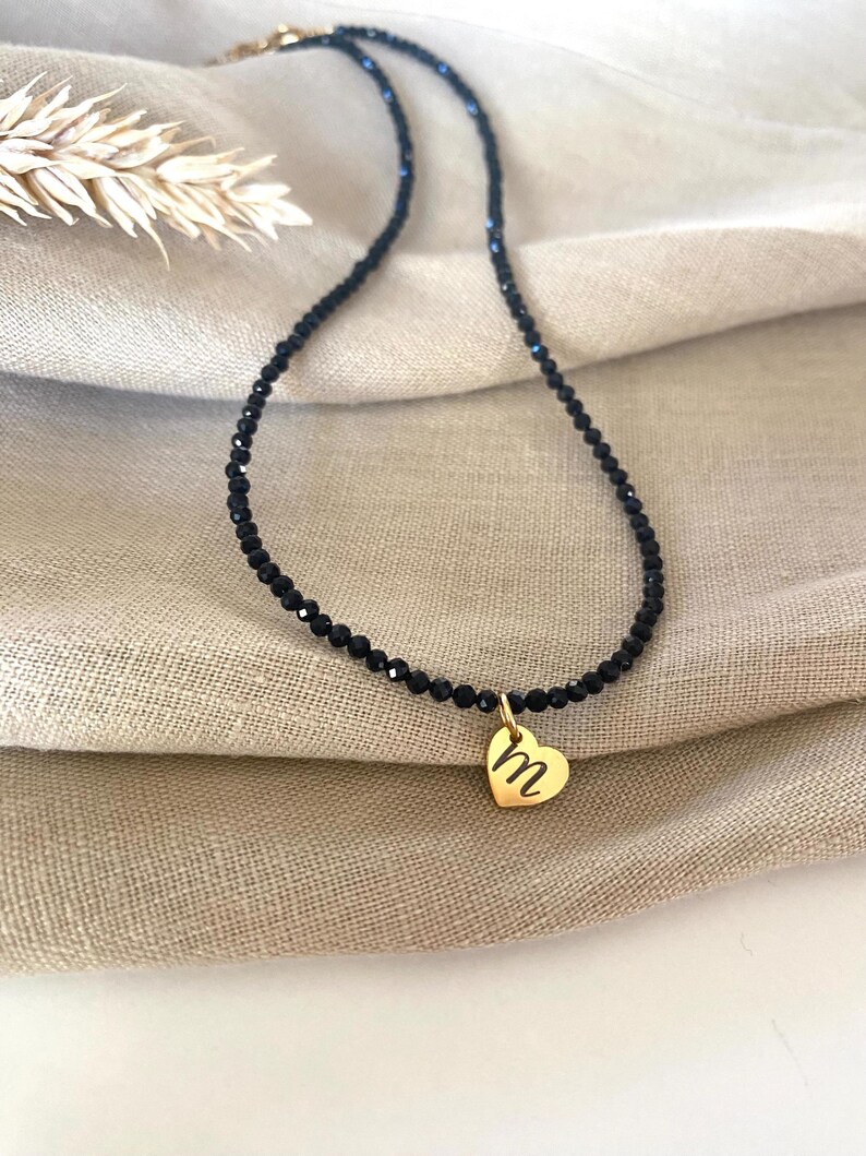 Puede incluir: Un delicado collar de cuentas negras con un colgante de coraz&oacute;n dorado. El colgante de coraz&oacute;n presenta la letra "m" en cursiva. El collar se muestra sobre una tela de tono neutro.