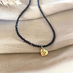 Puede incluir: Un collar de cuentas negras con un colgante de coraz&oacute;n dorado. El colgante presenta la letra "m" en un estilo manuscrito. El collar se muestra sobre un fondo de tela beige.