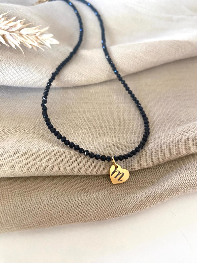 Puede incluir: Un collar de cuentas negras con un colgante de coraz&oacute;n dorado. El colgante presenta la letra "m" en un estilo manuscrito. El collar se muestra sobre un fondo de tela beige.