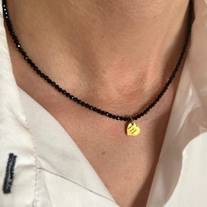 Puede incluir: Un delicado collar de cuentas negras con un peque&ntilde;o colgante de coraz&oacute;n dorado. El colgante de coraz&oacute;n tiene un dise&ntilde;o sutil y texturizado. El collar se lleva sobre una camisa blanca.