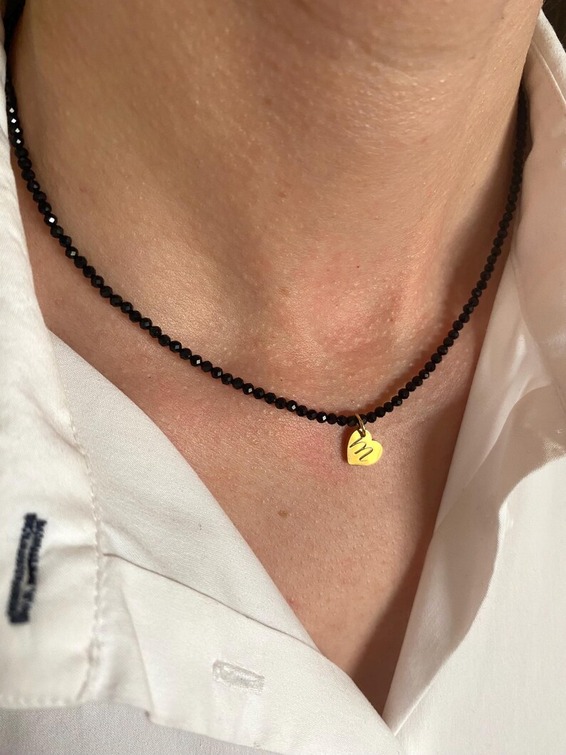 Puede incluir: Un delicado collar de cuentas negras con un peque&ntilde;o colgante de coraz&oacute;n dorado. El colgante de coraz&oacute;n tiene un dise&ntilde;o sutil y texturizado. El collar se lleva sobre una camisa blanca.