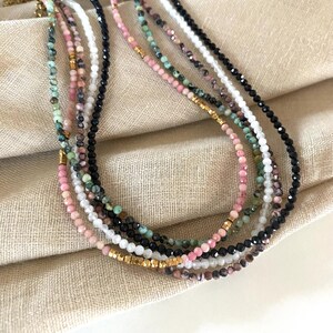 Puede incluir: Una colecci&oacute;n de delicados collares de cuentas en varios colores, incluyendo rosa, negro, blanco y verde. Los collares tienen peque&ntilde;as cuentas redondas y detalles dorados. Se exhiben sobre una superficie de tela beige.