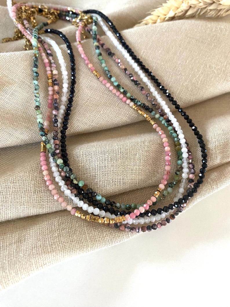 Puede incluir: Una colecci&oacute;n de delicados collares de cuentas en varios colores, incluyendo rosa, negro, blanco y verde. Los collares tienen peque&ntilde;as cuentas redondas y detalles dorados. Se exhiben sobre una superficie de tela beige.