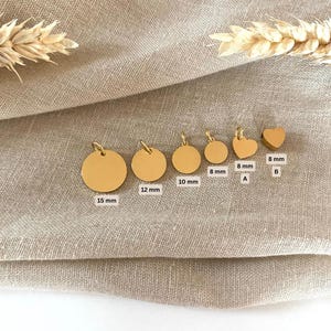 Puede incluir: Una colecci&oacute;n de colgantes dorados de varias formas y tama&ntilde;os. Los colgantes incluyen formas redondas y de coraz&oacute;n, con tama&ntilde;os que van desde 8 mm hasta 15 mm. Los colgantes se muestran sobre un fondo de tela de color neutro.