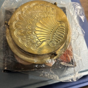 May include: A round brass coaster set featuring an embossed shell design on the top surface. The coasters are stacked together and enclosed in a clear plastic protective wrap, showing a metallic gold finish with detailed engraved lines.
