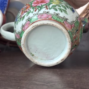 May include: A small, round, porcelain tea cup with a handle. The cup is decorated with a floral pattern in pink, red, green, and yellow. The base of the cup is a pale green color.