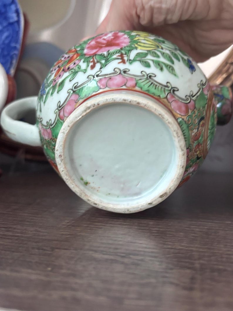 May include: A small, round, porcelain tea cup with a handle. The cup is decorated with a floral pattern in pink, red, green, and yellow. The base of the cup is a pale green color.