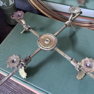 Peut inclure: Un support métallique en forme de croix doté de quatre bras réglables s'étendant à partir d'un moyeu central circulaire. Chaque bras se termine par un pied décoratif à bords festonnés. Le métal présente une finition vieillie et ternie.