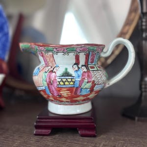 May include: A small, antique porcelain pitcher with a curved handle and a wide spout. The pitcher is decorated with a colorful scene of figures in traditional clothing. It sits on a small, dark red wooden stand.