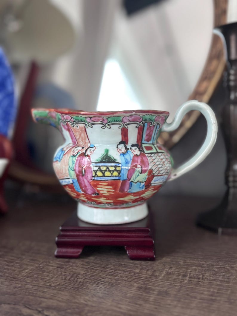May include: A small, antique porcelain pitcher with a curved handle and a wide spout. The pitcher is decorated with a colorful scene of figures in traditional clothing. It sits on a small, dark red wooden stand.