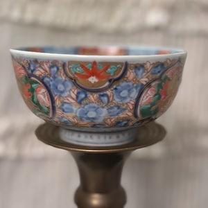 Puede incluir: Un cuenco de cerámica decorativo con un patrón floral en azul, rojo, verde y dorado. El cuenco está sostenido por un soporte de latón, creando una exhibición elegante. El diseño del cuenco presenta detalles intrincados.