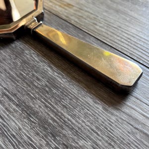 May include: A vintage hand mirror with a rectangular, beveled mirror and a gold-toned handle. The handle has a flat, elongated shape. The mirror's frame and handle show signs of wear, suggesting its age. The mirror is resting on a wooden surface.