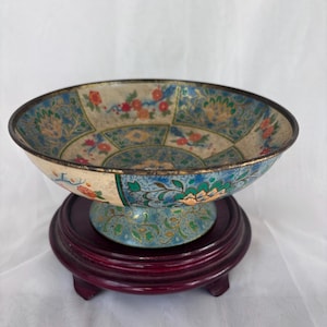 May include: A decorative bowl with a floral design in shades of blue, orange, and green, set on a dark red wooden stand. The bowl has a gold rim and a detailed pattern.