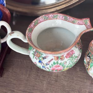 May include: A small, antique porcelain creamer with a white handle and spout. The creamer features a floral and bird pattern in pink, green, and blue. The rim is trimmed with a pink and green floral design. The creamer is on a wooden surface.