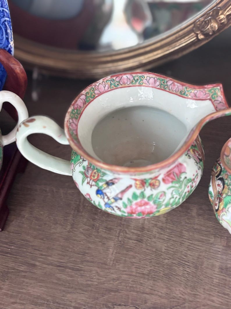 May include: A small, antique porcelain creamer with a white handle and spout. The creamer features a floral and bird pattern in pink, green, and blue. The rim is trimmed with a pink and green floral design. The creamer is on a wooden surface.