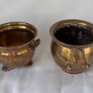 May include: Two antique brass planters. The planter on the left has a round shape with three small feet and two handles. The planter on the right has a round shape with a rope design around the top. Both planters have a golden color.