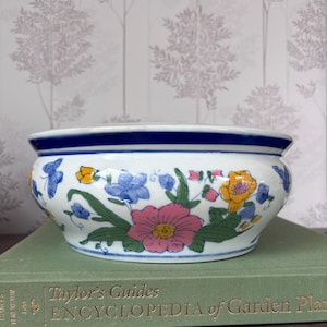 Peut inclure: Un bol en céramique blanc avec un bord bleu et un motif floral. Le bol présente des fleurs roses, jaunes et bleues avec des feuilles vertes et des papillons bleus. Le bol est posé sur un livre vert.