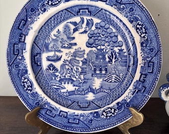 May include: A blue and white ceramic plate with a Willow pattern design. The plate features a detailed landscape scene with pagodas, bridges, and birds. The plate is displayed on a gold-colored stand.