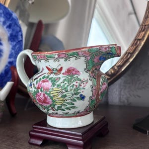May include: A small, antique porcelain creamer with a floral design in pink, green, yellow, and blue. The creamer has a white base and handle, and sits on a dark red wooden stand. A butterfly is visible on the side.