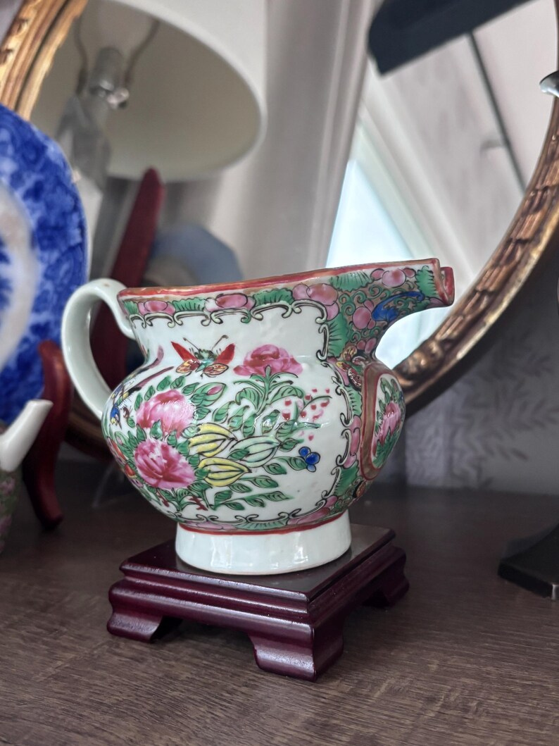 May include: A small, antique porcelain creamer with a floral design in pink, green, yellow, and blue. The creamer has a white base and handle, and sits on a dark red wooden stand. A butterfly is visible on the side.