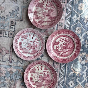 May include: Four antique pink and white ceramic plates with a classic Willow pattern. Each plate features a detailed landscape scene with pagodas, trees, and bridges. The plates are arranged on a patterned rug.