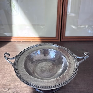 May include: A silver-toned bowl with a hammered texture and decorative rim. The bowl has two ornate handles and sits on a dark, round base. The background includes framed artwork.