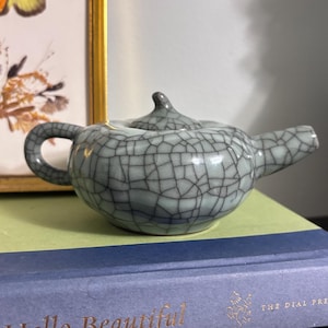 Könnte beinhalten: Eine kleine, hellgrüne Keramik-Teekanne mit einem Craquelé-Glasurmuster. Die Teekanne hat einen gebogenen Griff, einen Ausguss und einen Deckel mit einem kleinen Knopf. Die Teekanne liegt auf einem grünen und blauen Buch mit den Worten "Hello Beautiful" in goldfarbenen Buchstaben.