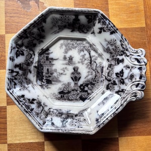 May include: An octagonal, antique-style plate with a black and white Asian-inspired landscape design. The plate features a detailed floral border and a central scene with pagodas and trees. The plate has a decorative handle.