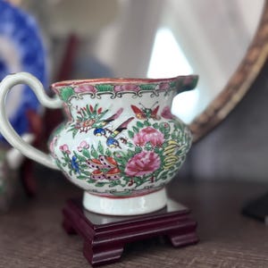 May include: A small, ornate porcelain creamer with a white base, adorned with colorful floral and bird designs. The creamer has a curved handle and a spout, and it sits on a small, dark red wooden stand. The overall design is a classic, vintage style.