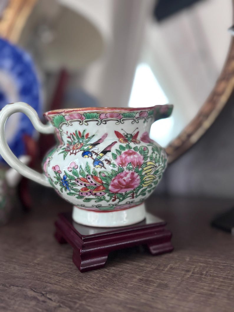 May include: A small, ornate porcelain creamer with a white base, adorned with colorful floral and bird designs. The creamer has a curved handle and a spout, and it sits on a small, dark red wooden stand. The overall design is a classic, vintage style.