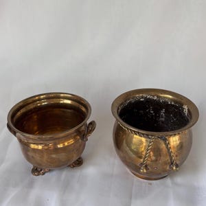 May include: Two antique brass planters. The planter on the left has three small feet and two handles. The planter on the right has a rope design around the top. Both planters have a shiny, golden finish and are empty.